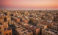 A surrealistic image depicts the bustling cityscape of Alexandria, Egypt, with a lone crowed scene in the background The sky is ablaze with orange and pink hues, casting a warm glow on the buildings and the surrounding buildings The camera focuses on the crowded scene